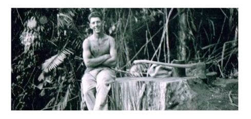 Ted Simms in the bush, seated beside a felled log and a broad axe.