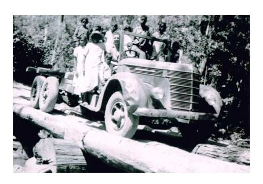 A loaded timber truck crossing a bridge of felled logs, with a group of people riding on the running boards and tray.